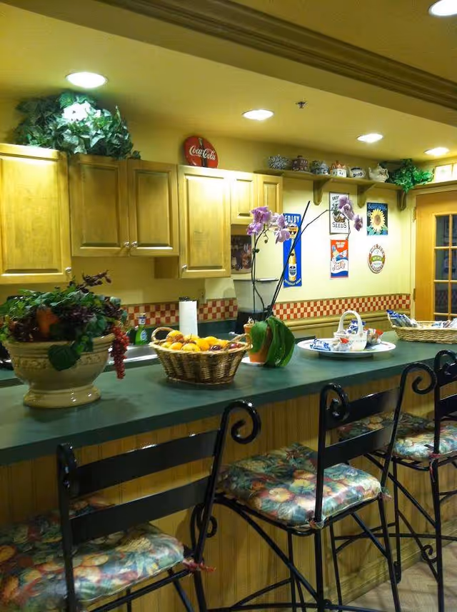 A kitchen area with a green countertop and wooden cabinets. There are three high chairs with floral-patterned cushions along the counter. On the counter, there is a basket of fruit, a potted plant, a paper towel roll, and a tray with cups and a teapot. The wall behind the counter has decorative signs and shelves with small decorative items. The lighting is warm with recessed ceiling lights.