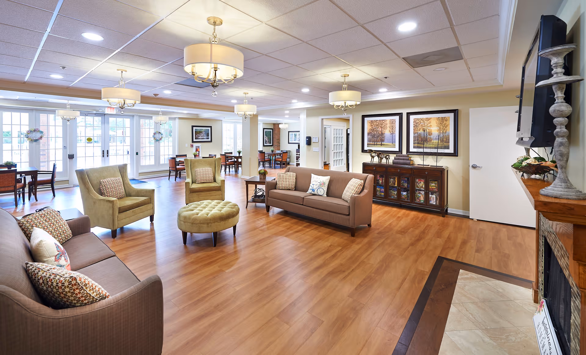A spacious and well-lit living room area in a senior living facility with wooden flooring, two brown sofas, two green armchairs, a round green ottoman, and several tables and chairs near large windows and glass doors. The room features ceiling lights, framed artwork on the walls, a wooden cabinet with decorative items, and a fireplace with a TV mounted above it.