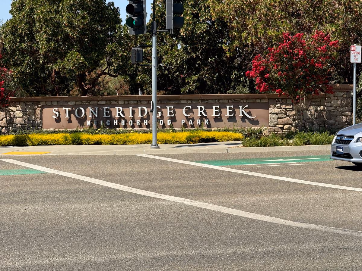Stone entrance sign reading "Stoneridge Creek Neighborhood Park" beside a roadway with landscaping, a traffic signal pole, and a partially visible car.