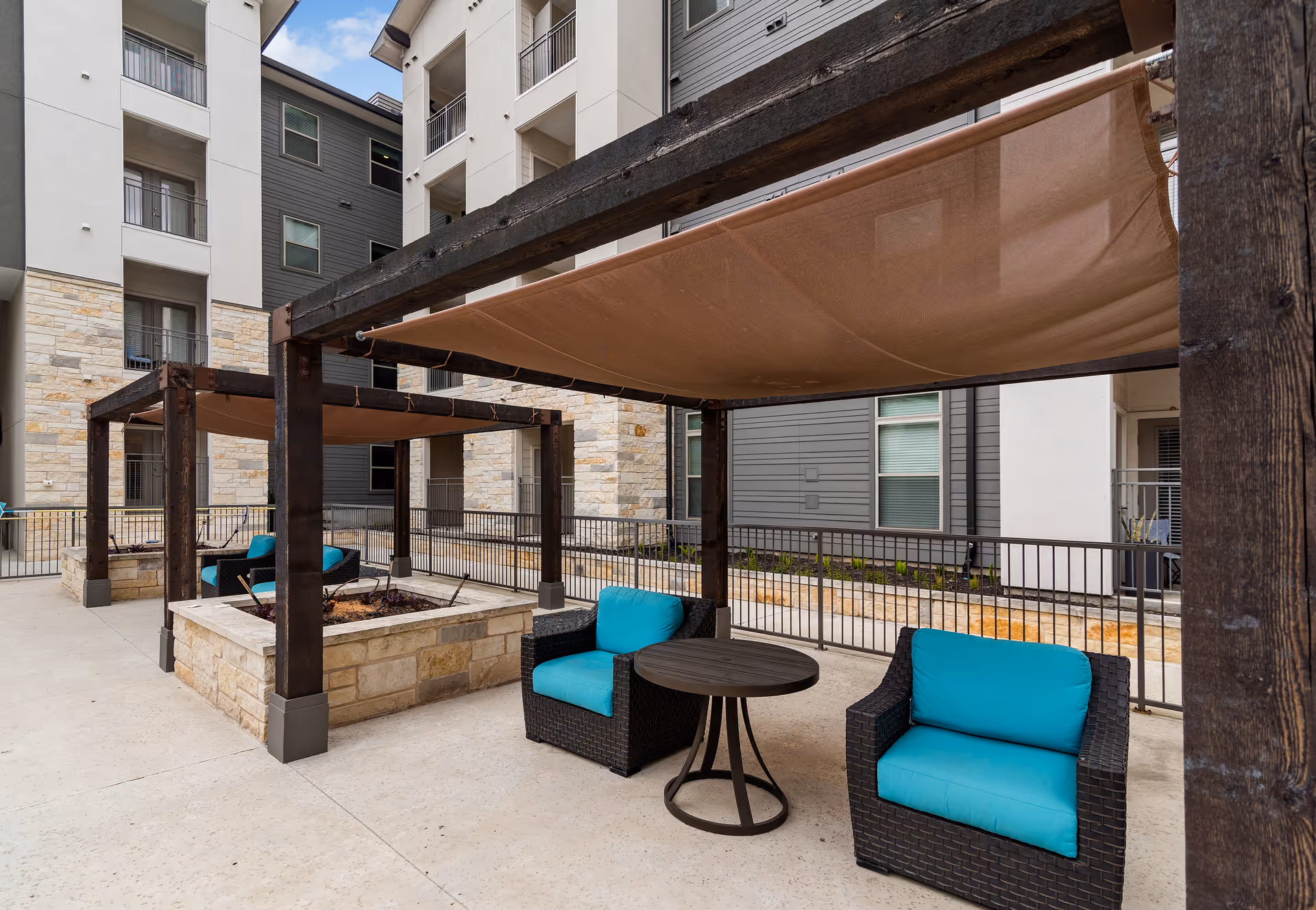Outdoor patio area at ArborView Circle C Ranch featuring two black wicker chairs with bright turquoise cushions and a small round black table between them. The seating area is shaded by a brown fabric canopy supported by dark wooden beams. The patio is surrounded by a multi-story building with stone and gray siding exterior walls and several windows and balconies.