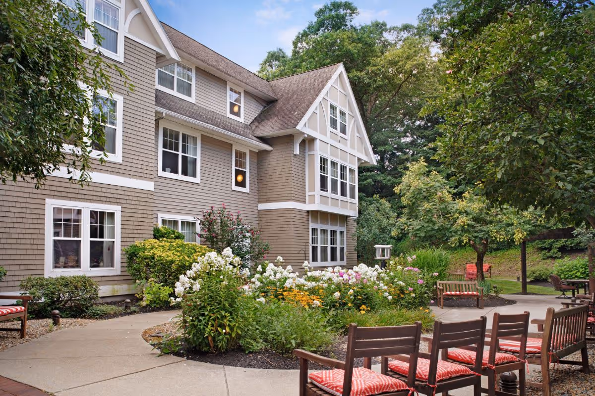 Outdoor garden area at Sunrise of Norwood featuring a paved walkway, flower beds with white and yellow flowers, several wooden benches with red patterned cushions, and a multi-story building with beige siding and white trim in the background surrounded by trees and greenery.
