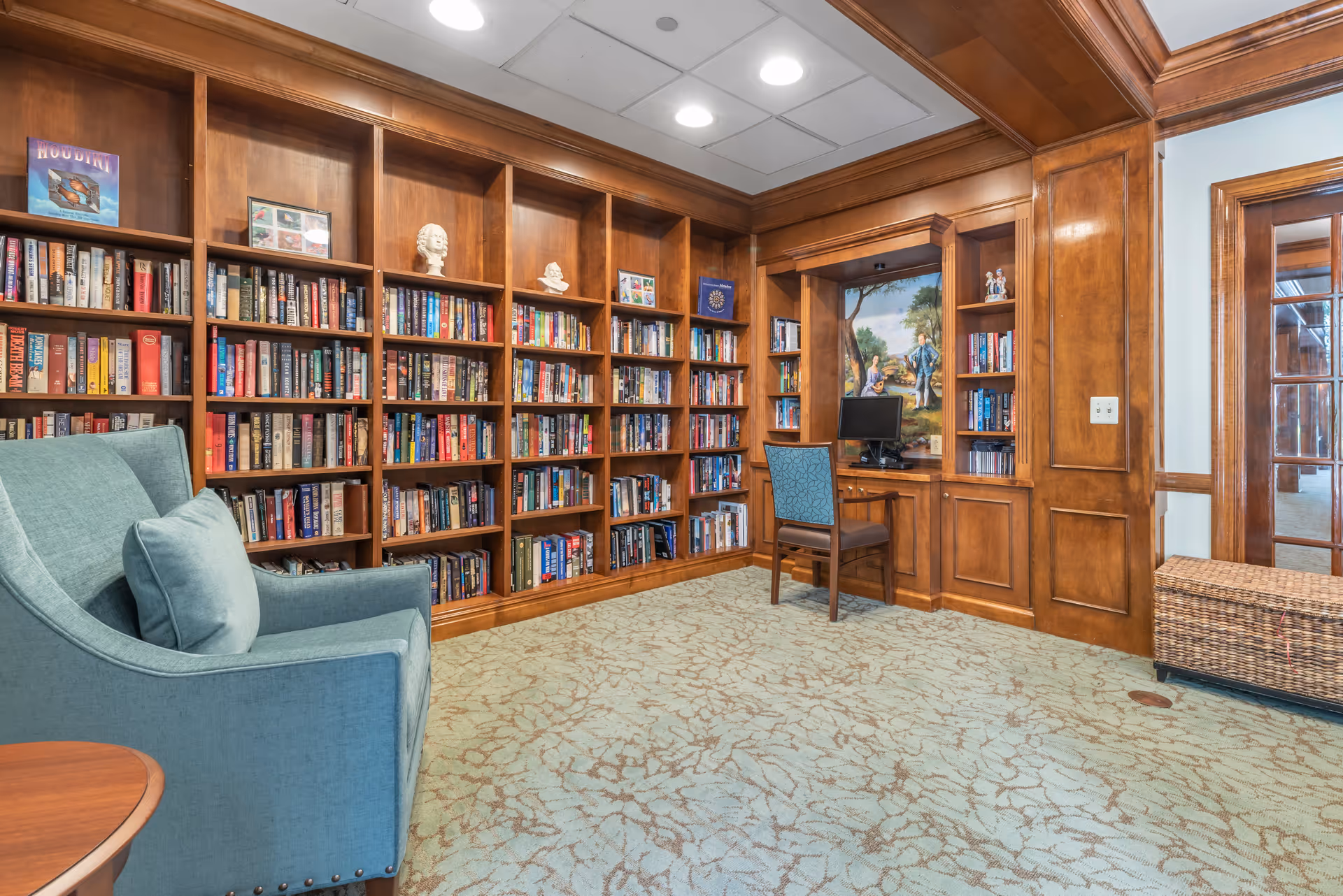 A cozy library room with wooden bookshelves filled with books and decorative busts. There is a blue upholstered armchair with a matching pillow on the left side and a wooden desk with a computer and a blue patterned chair on the right. The room has a patterned carpet and wood-paneled walls with a painting behind the desk.