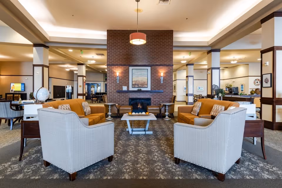 A spacious and well-lit senior living facility common area featuring two orange sofas and two beige armchairs arranged around a modern coffee table with decorative items. A brick fireplace with a framed picture above it is centered on the far wall, flanked by two wall sconces. The room has a patterned carpet, multiple columns, and additional seating and tables in the background.