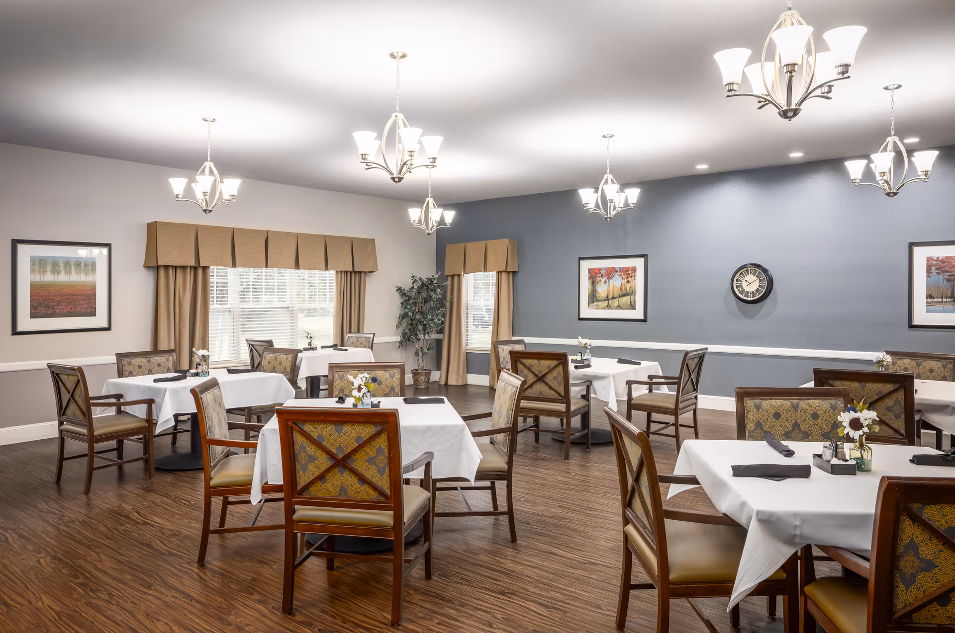 A dining room with multiple square tables covered with white tablecloths, each set with black napkins and small floral centerpieces. The room has wooden chairs with patterned upholstery, wood flooring, beige and blue walls, framed artwork, a clock, and several ceiling light fixtures.
