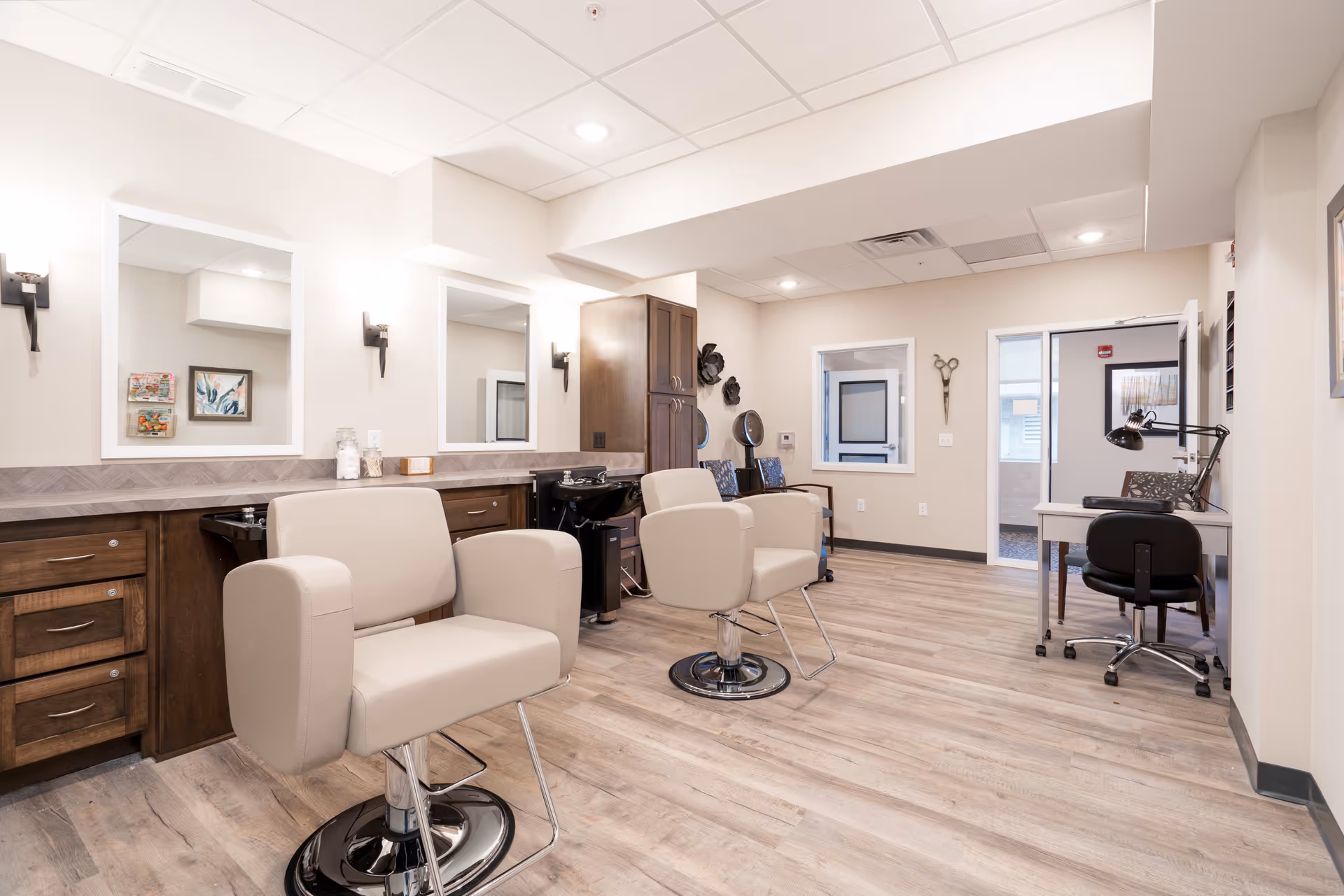 Interior view of a senior living facility's salon area with two beige salon chairs in front of mirrors and sinks, wooden cabinets, and a small desk with a black office chair. The room has light-colored walls and wood flooring.
