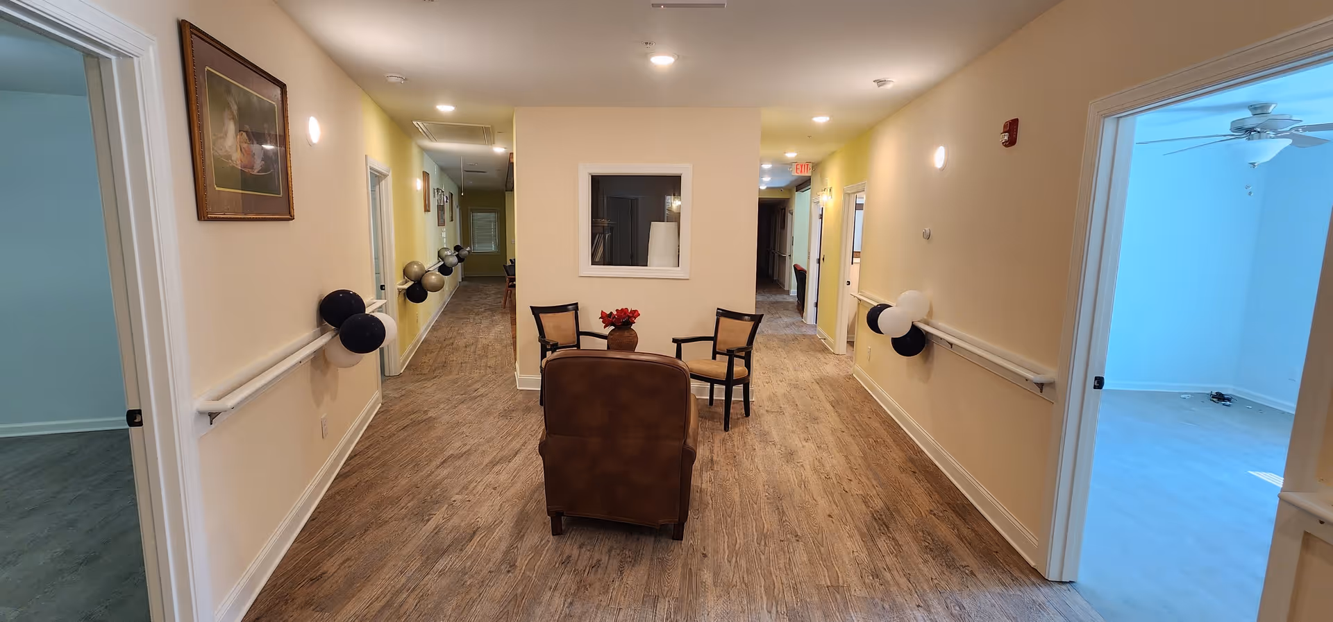 A hallway in Green Park Senior Care Home with wood flooring and beige walls. The hallway has handrails on both sides decorated with black and white balloons. There are three chairs and a small table with a flower vase placed in the center near the end of the hallway. Doors lead to other rooms on both sides, and the area is well-lit with ceiling lights.
