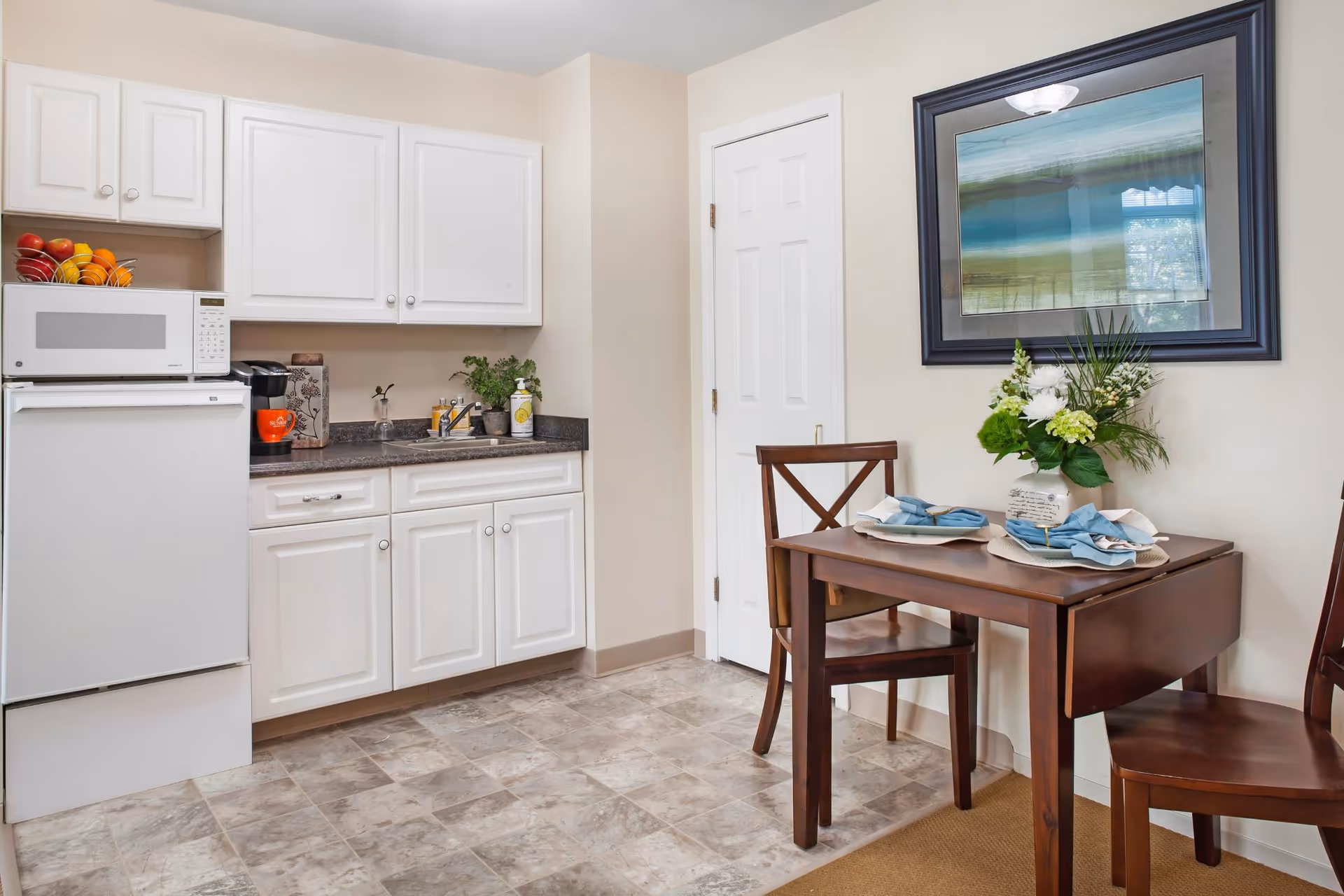 A small kitchen area with white cabinets, a white microwave on top of a white mini refrigerator, a coffee maker, and a sink with a few items on the counter. Adjacent to the kitchen is a small wooden dining table set for two with blue napkins and a floral centerpiece. A framed picture hangs on the wall above the table.