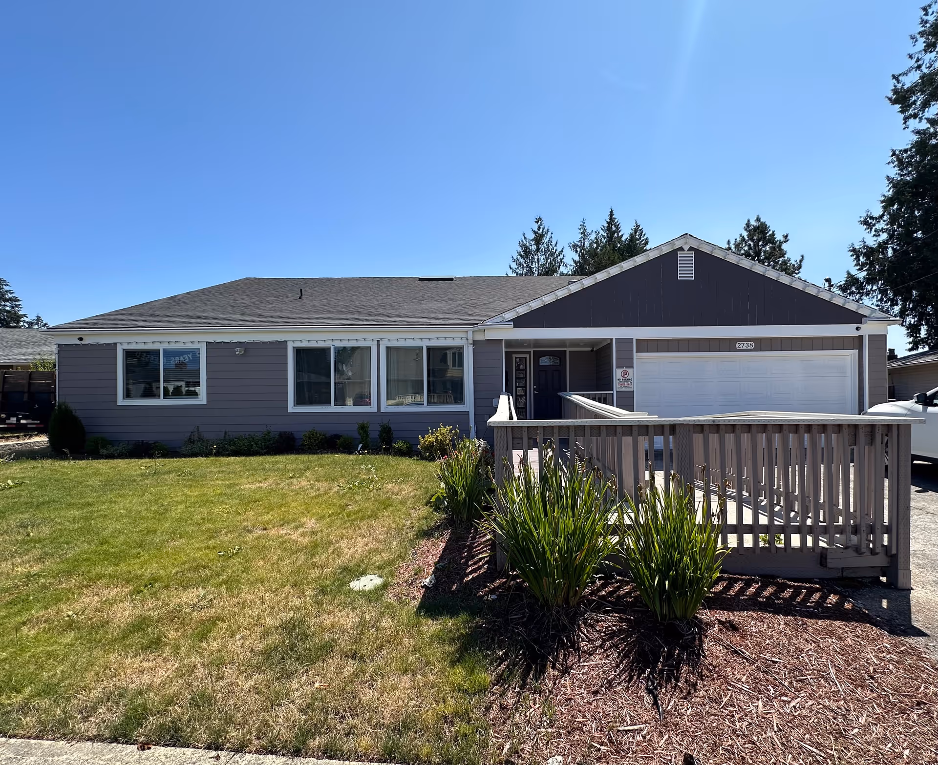 Single-story residential building with gray siding and a dark gray roof under a clear blue sky. The front yard has a green lawn and some plants near a wooden ramp leading to the front door. There is a two-car garage on the right side of the house.