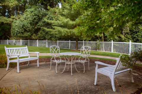 Outdoor patio area with white metal table and two chairs, flanked by two white wooden benches, surrounded by green grass, trees, and a white fence in the background.