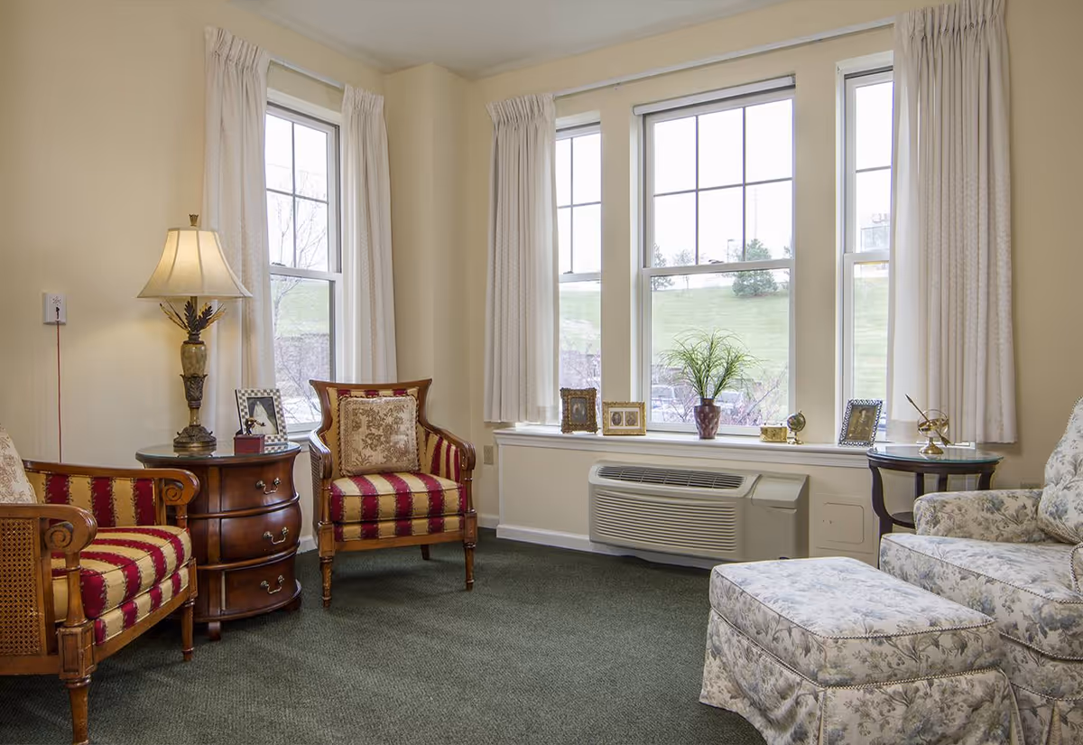 A cozy living room area with two wooden armchairs upholstered in red and gold striped fabric, a floral patterned armchair with matching ottoman, a wooden side table with a lamp, and a window sill decorated with framed photos and a potted plant. The room has large windows with cream-colored curtains and green carpet flooring.