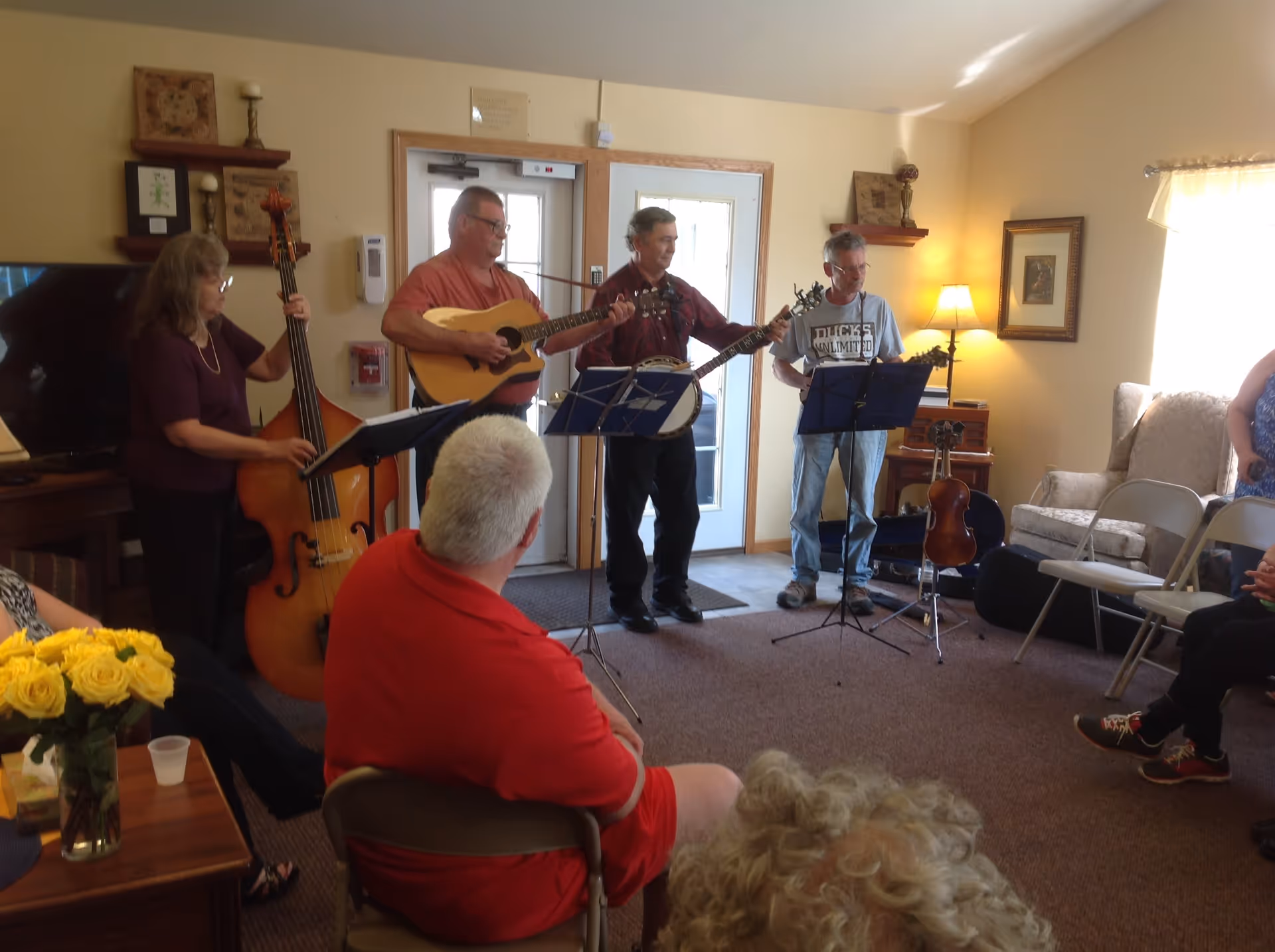 Musicians playing guitar, banjo and upright bass in a cozy living-room style common area while residents sit and listen.