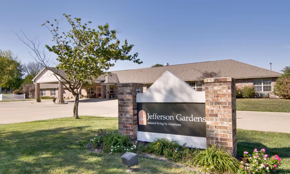 Sign reading 'Jefferson Gardens' in front of a single-story brick senior living building with driveway, lawn, and trees.