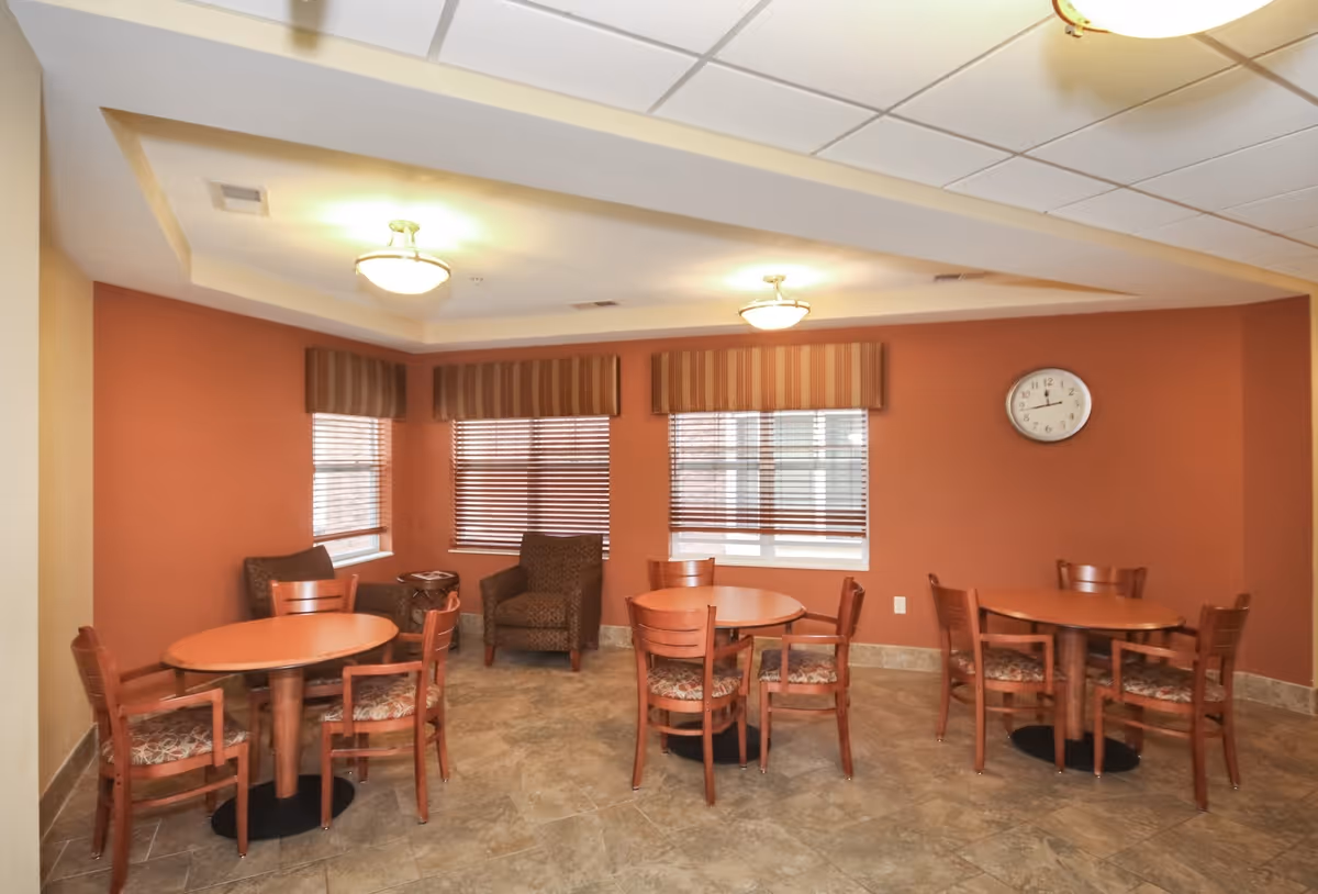 A common area with three round wooden tables, each surrounded by four wooden chairs with patterned cushions. Two armchairs and a small side table are placed near the windows with striped valances. The walls are painted a warm reddish color, and a clock is mounted on the wall. The floor is tiled, and ceiling lights provide illumination.