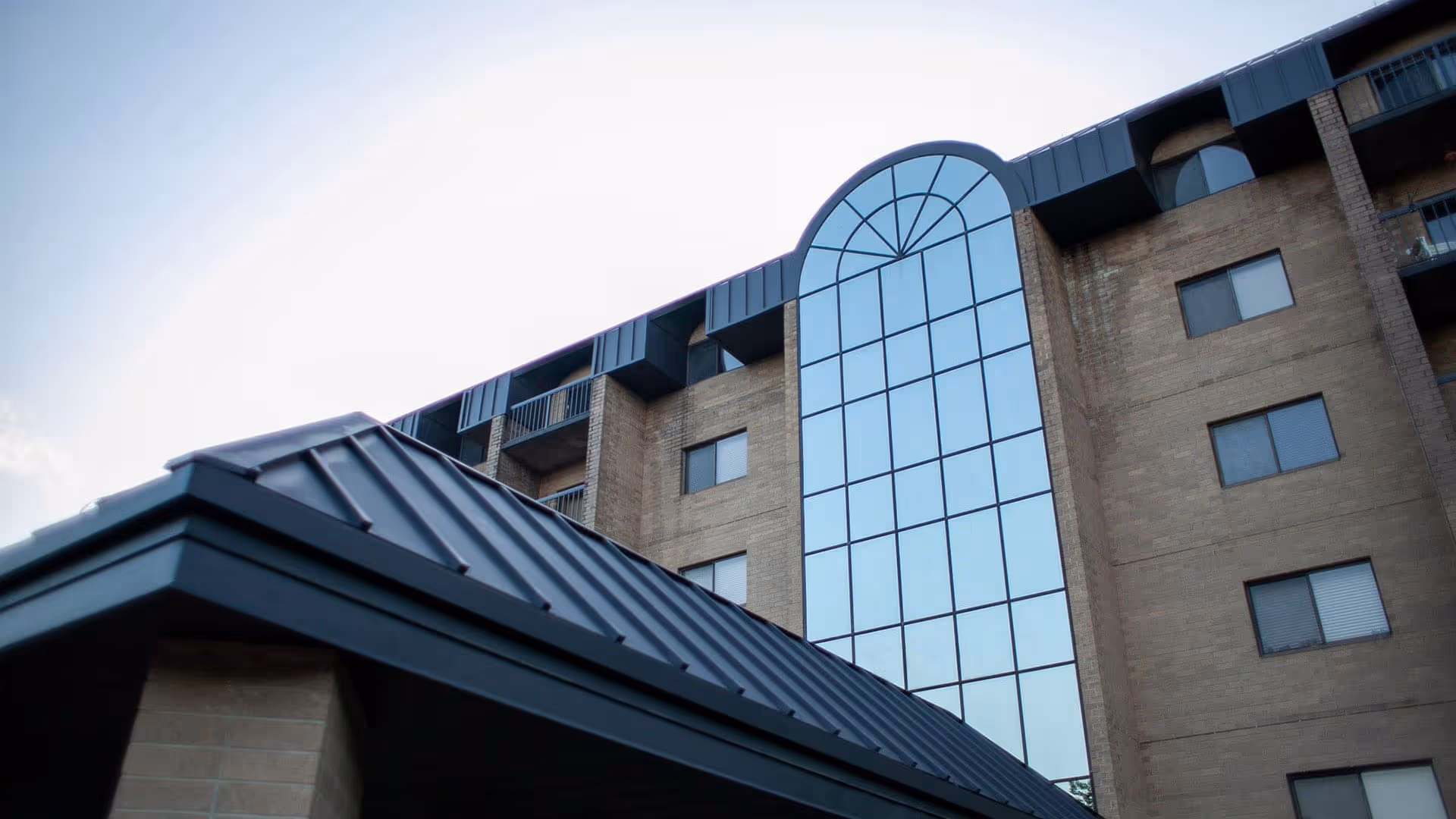 Low-angle view of a multi-story brick building facade with a tall arched glass atrium and a dark metal roof.