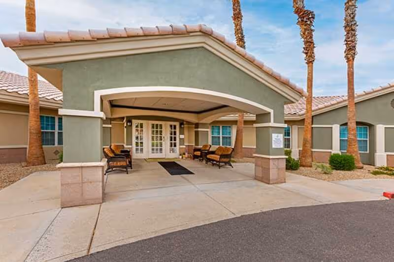 Entrance of a senior living facility with a covered driveway, outdoor seating with chairs and small tables, palm trees, and a clear sky.