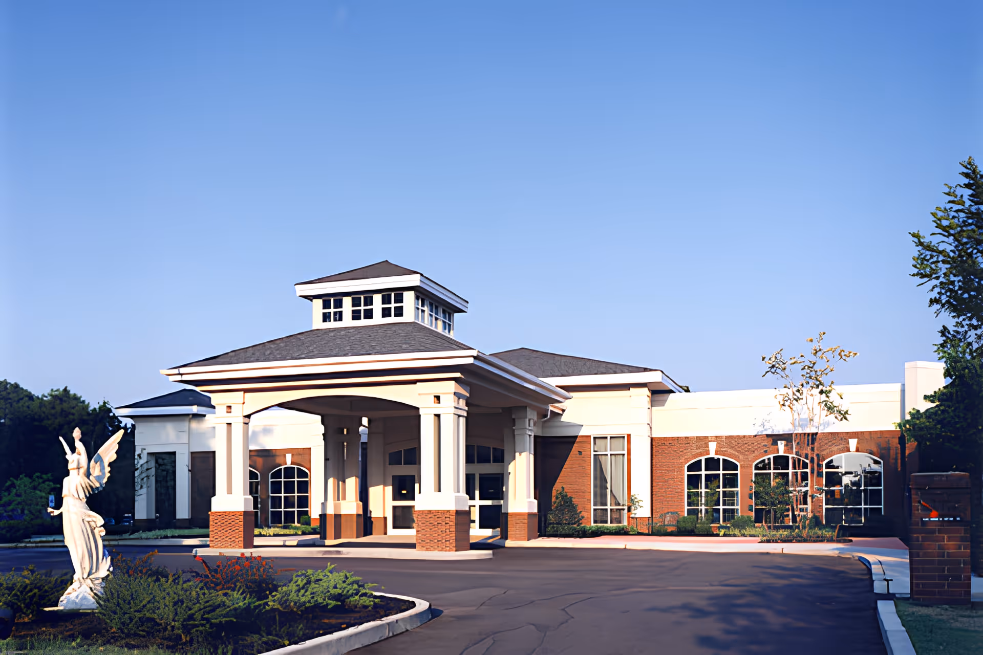 Exterior view of a senior living facility named Ave Maria with a covered entrance supported by columns, large windows, and a statue of an angel in the landscaped front area under a clear blue sky.