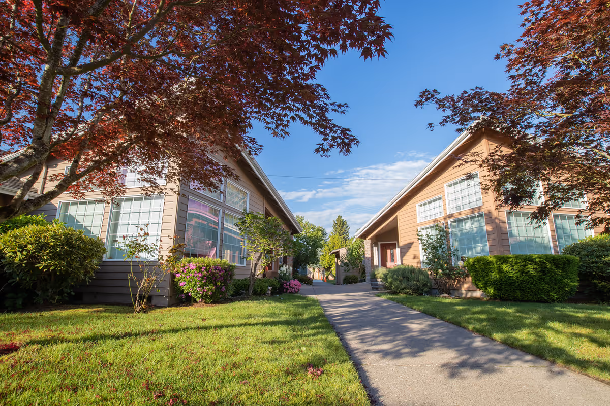 A sunny outdoor pathway between two single-story buildings with large windows, surrounded by green grass, bushes, and trees with red leaves under a clear blue sky.