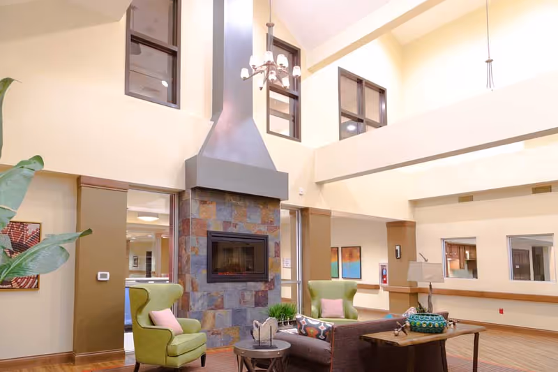 A bright and spacious living room area with high ceilings and large windows. The room features a modern stone fireplace with a metal chimney, two green armchairs with pink cushions, a brown sofa with colorful pillows, a wooden coffee table with decorative items, and a side table with a lamp. The walls are painted cream with brown accents, and there are framed artworks and a plant visible.