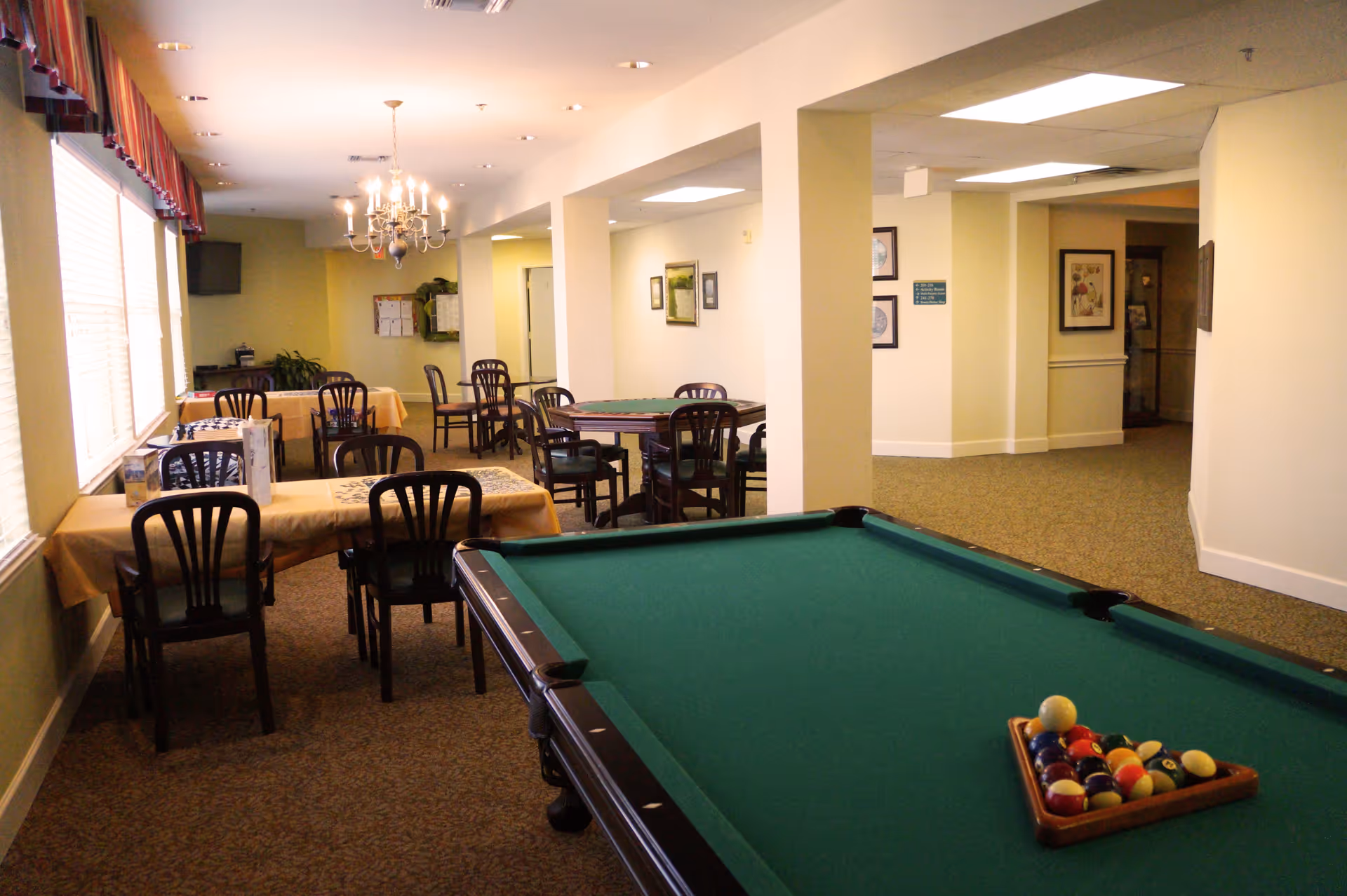 Interior room with a pool table in the foreground and several tables with chairs covered in yellow tablecloths in the background. The room has large windows with blinds and a chandelier hanging from the ceiling. The walls are light-colored with framed pictures and a carpeted floor.