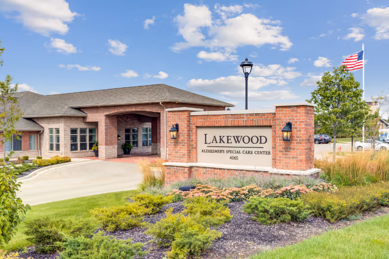 Exterior view of Lakewood Alzheimer's Special Care Center building with a brick entrance sign, landscaped greenery, an American flag, and a clear blue sky with scattered clouds.