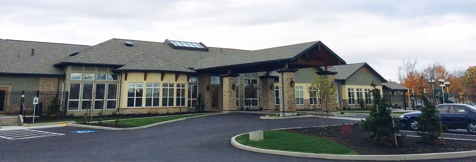 Front entrance of a single-story memory care building with large windows, a covered porte-cochère, and a parking area.