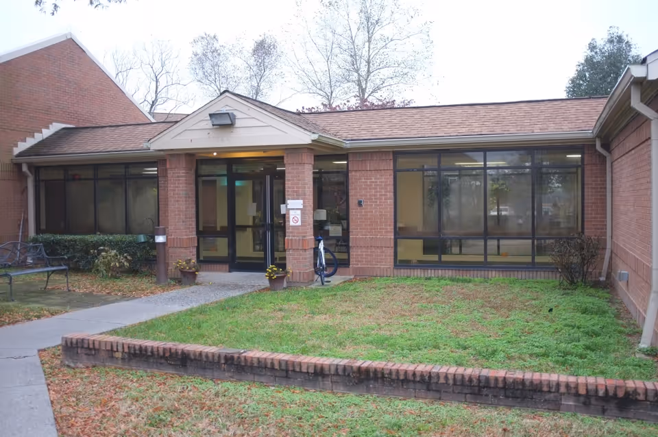 Exterior view of a brick building with large windows and a glass door entrance. There is a small grassy area with a low brick border in front, a concrete pathway leading to the entrance, and a bench on the left side near some bushes.