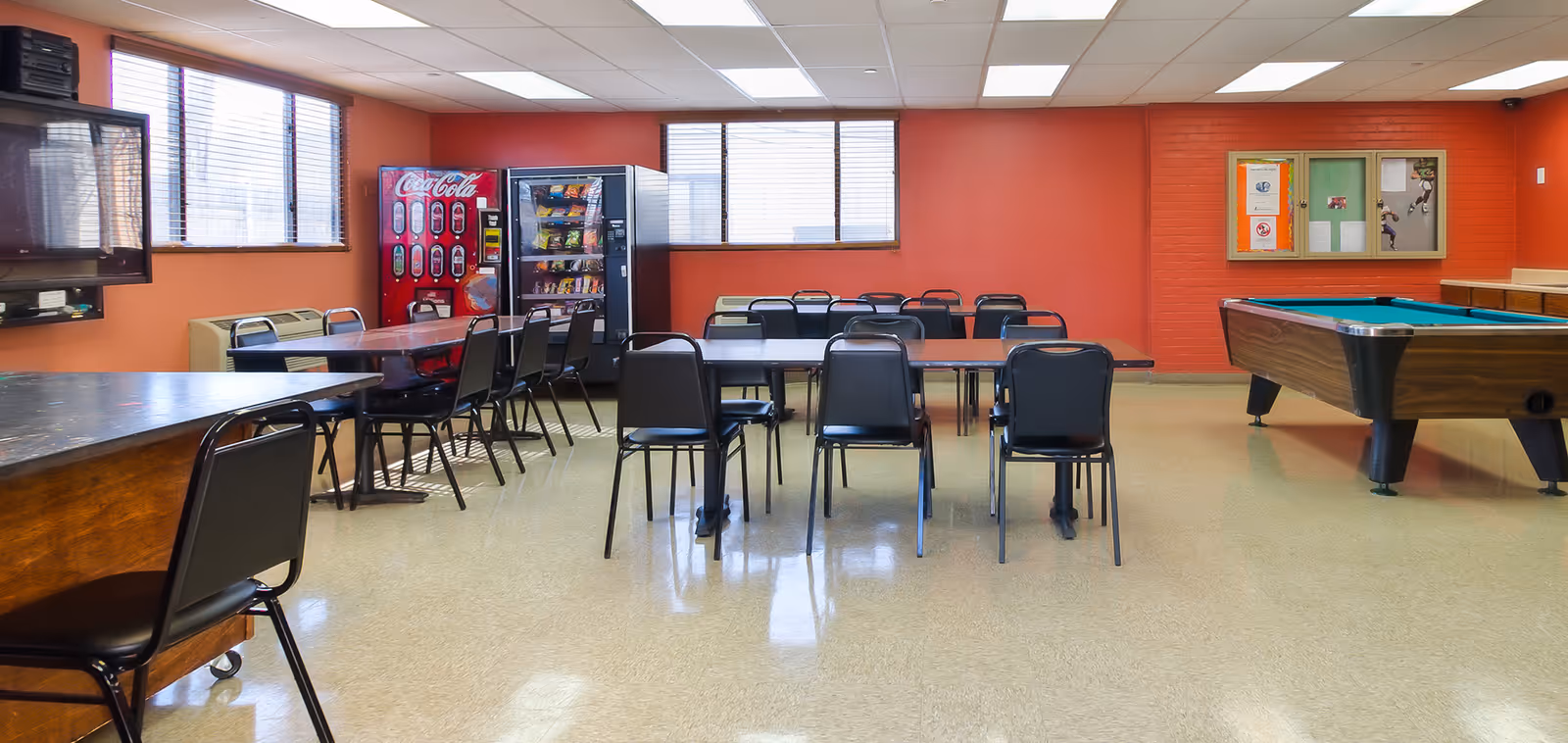A community room with several tables and black chairs arranged in rows. There is a Coca-Cola vending machine and a snack vending machine against the far wall with windows above them. On the right side of the room, there is a pool table. The walls are painted orange, and there is a bulletin board on the right wall.