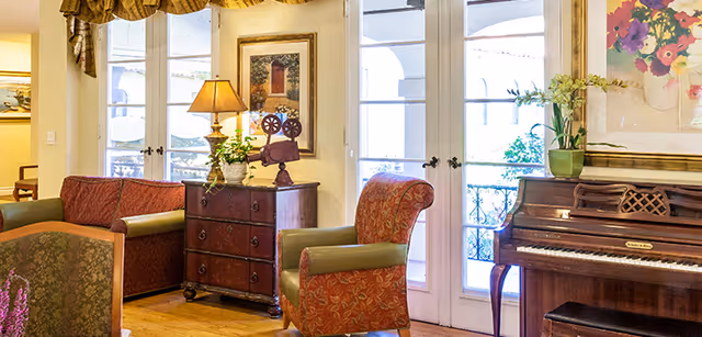 A cozy living room area in Foothill Retirement featuring a wooden floor, a red patterned armchair, a green armchair, a wooden chest of drawers with a lamp and decorative items, a piano with a floral painting above it, and French doors leading outside.