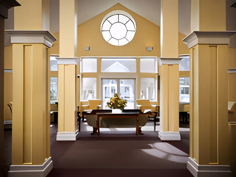Interior view of a spacious room with tall beige columns and a large circular window above a set of rectangular windows. In the center, there is a wooden table with a flower arrangement, surrounded by chairs. The room has a high ceiling and carpeted floor.