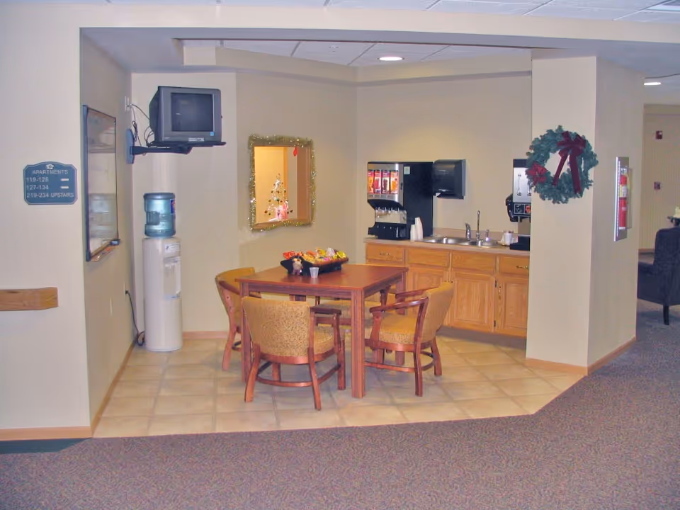 A small common area in a retirement community featuring a wooden table with four cushioned chairs, a water cooler, a wall-mounted TV, a counter with a beverage dispenser, a sink, and a paper towel dispenser. The walls are decorated with a wreath and a mirror with holiday decorations visible through the mirror.