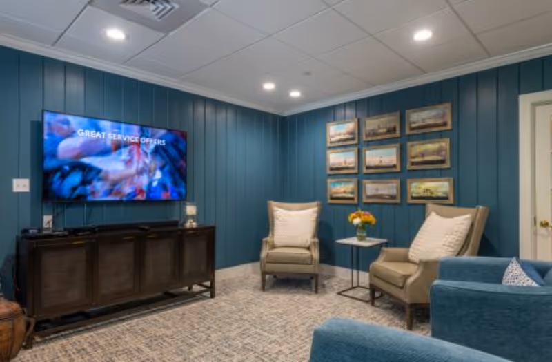 A cozy living room with teal paneled walls, a mounted flat-screen TV displaying the text 'GREAT SERVICE OFFERS,' two beige armchairs with white pillows, a small side table with a vase of flowers, a blue sofa, and a series of framed pictures on the wall.