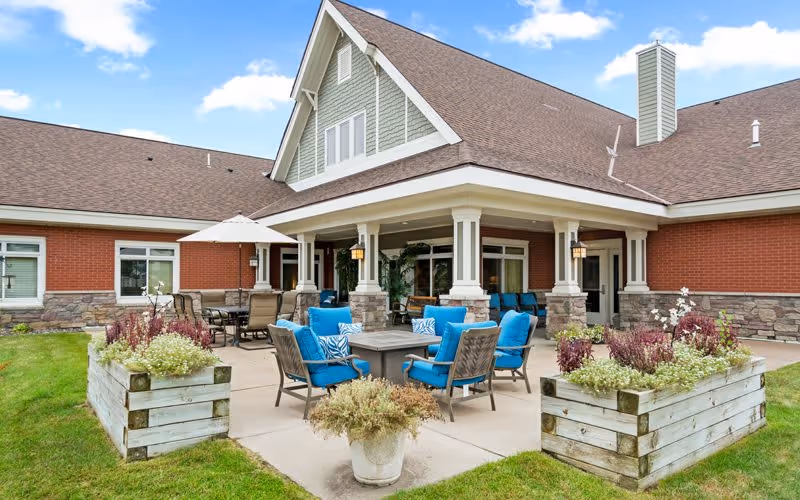 Outdoor patio area at Encore Assisted Living and Memory Care at North Branch featuring cushioned blue chairs around a fire pit table, additional seating under a large umbrella, and raised wooden planters with flowers and greenery. The building exterior has brick and stone accents with a covered porch and a peaked roof under a partly cloudy sky.