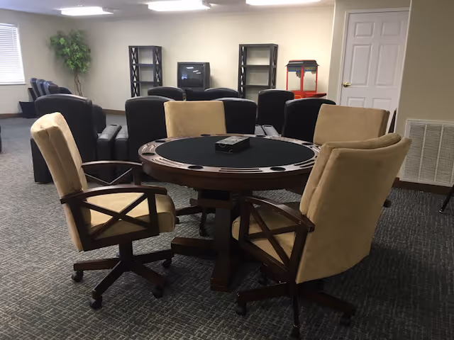 A senior living facility common room with a round poker table surrounded by four beige cushioned chairs on wheels. In the background, there are several black armchairs arranged facing a TV on a stand, with empty shelving units on either side. A popcorn machine is visible near a closed white door, and a potted plant is in the corner near a window.