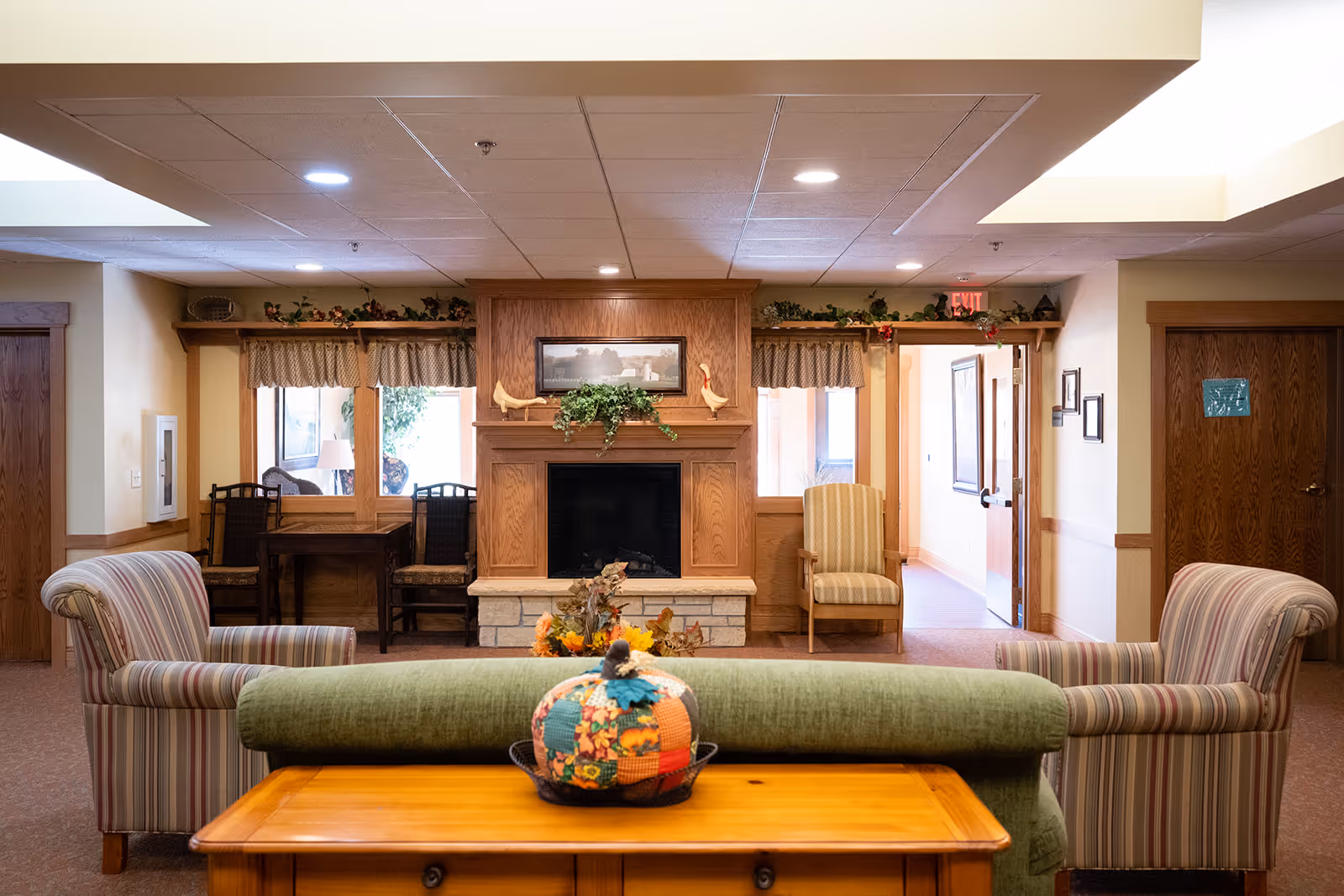 A cozy living room area in a senior living facility featuring a green couch with a decorative pumpkin on a wooden table behind it, two striped armchairs facing the couch, a wooden fireplace with decorative items and a framed picture above it, and a small seating area with chairs and a table near windows with curtains.