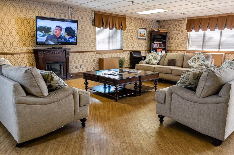 A cozy living room with beige sofas arranged around a dark wooden coffee table. The room features patterned wallpaper, two windows with brown valances, a flat-screen TV mounted above a decorative fireplace, and a bookshelf filled with books and games in the corner.