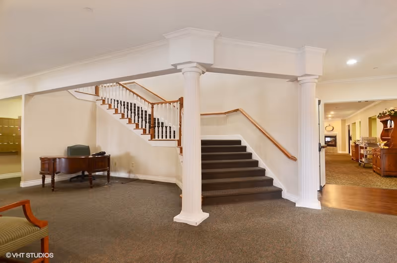 Interior view of a senior living facility lobby area with a staircase featuring white railings and wooden handrails. There are two white decorative columns supporting a beam above the stairs. To the left, there is a wooden desk with a green chair. The floor is carpeted, and the walls are painted light beige. In the background, a hallway with additional furniture and lighting is visible.