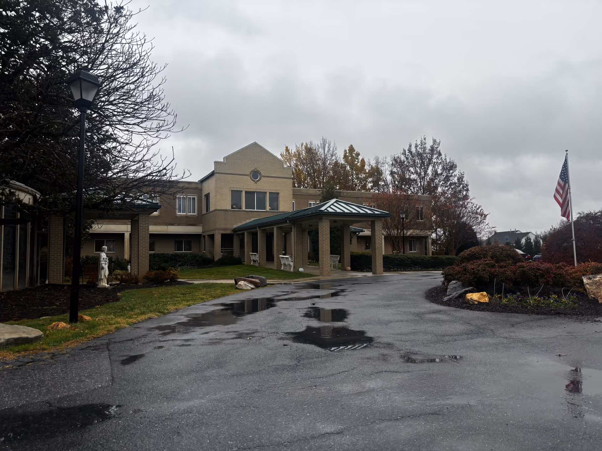 Exterior view of a senior living facility building on a cloudy day with wet pavement. The building has a covered entrance supported by columns, surrounded by trees and bushes, with an American flag on a flagpole to the right.