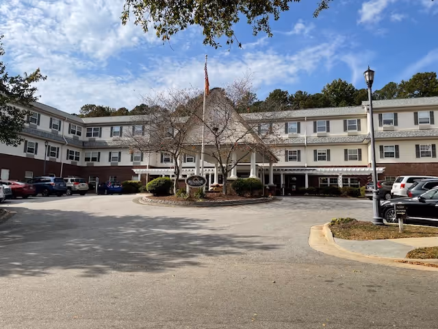 Front entrance of a three-story senior living building with a covered porte-cochere, circular driveway, flagpole, and parked cars.