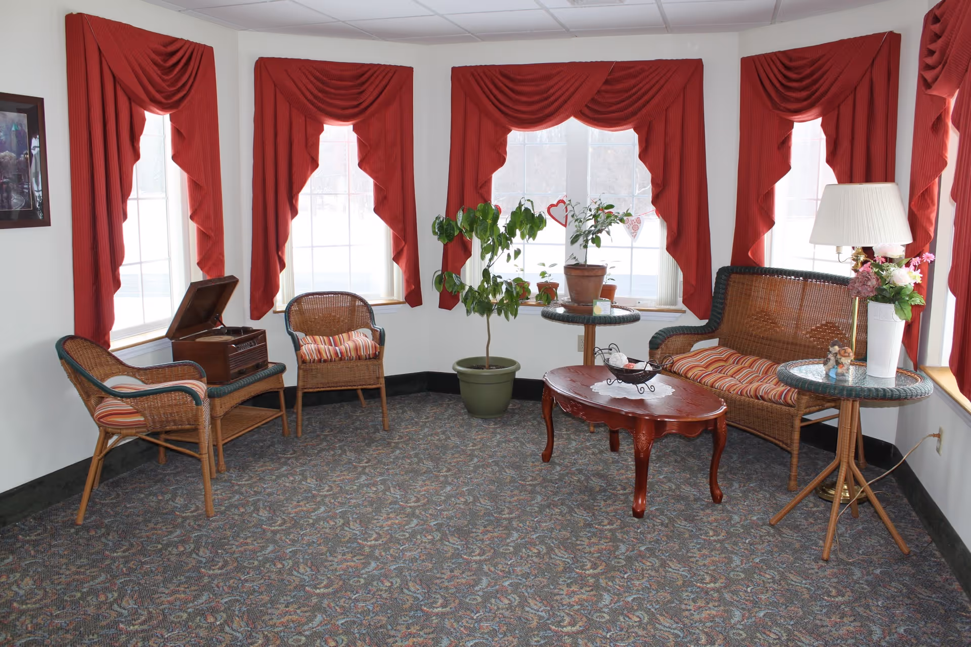 A cozy sitting area with wicker chairs and a loveseat arranged around a wooden coffee table beneath bay windows dressed with red drapes.
