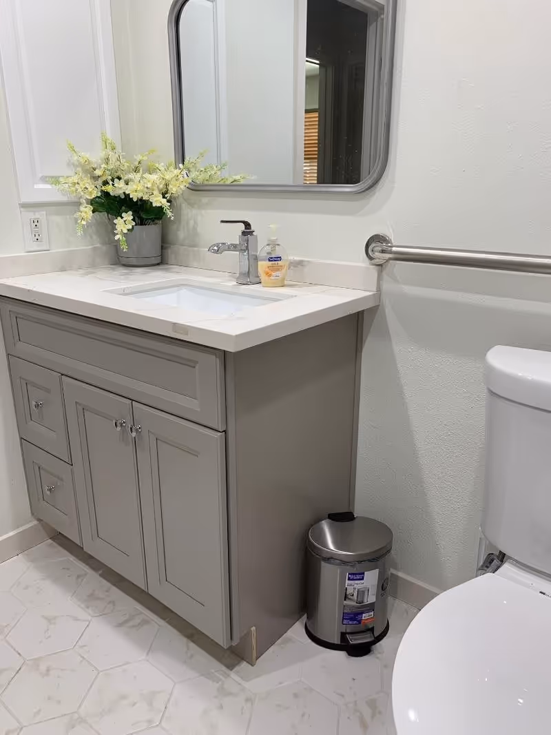 Bathroom vanity with a sink, mirror, floral arrangement, grab bar, small trash can, and a partially visible toilet.