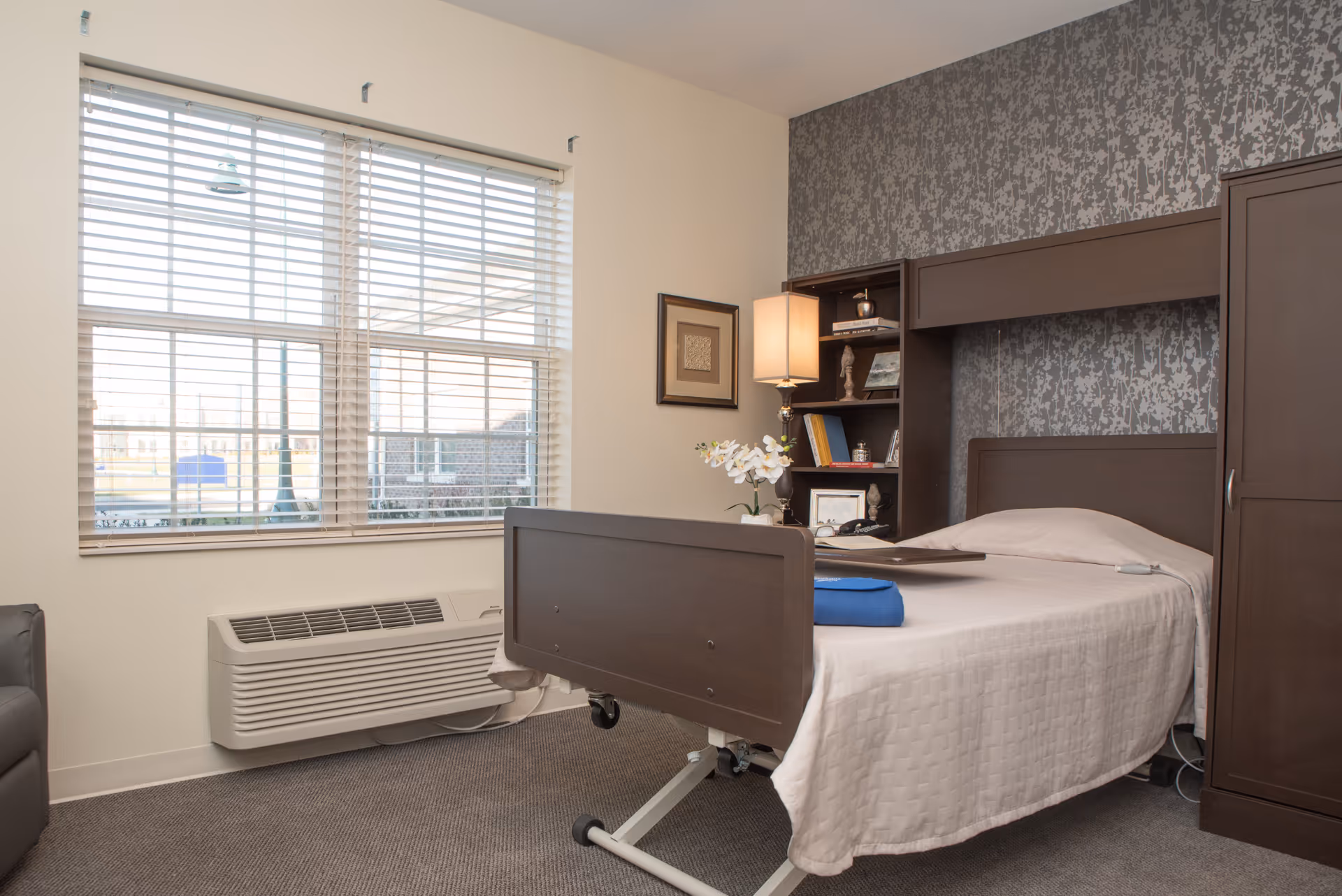 A neatly arranged bedroom in a senior living facility with a single hospital-style bed covered with a white quilt. The bed has a dark wooden headboard and footboard, and there is a blue towel folded on the bed. Next to the bed is a dark wooden shelving unit with books, decorative items, and a lamp. A window with white blinds allows natural light into the room, and a framed picture hangs on the wall. The floor is carpeted in a neutral tone.