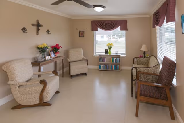 A bright communal sitting room with upholstered chairs, a small table with flowers, and a bookcase under the window.