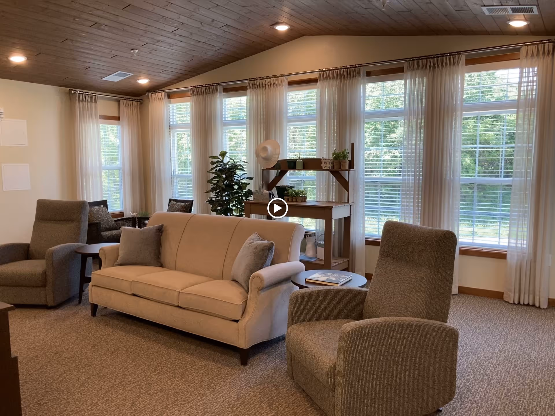 A cozy living room area with a beige sofa, two gray armchairs, a small round side table with a book, and a wooden shelf with potted plants and a hat. Large windows with white curtains allow natural light to fill the room, and the ceiling is wooden with recessed lighting.