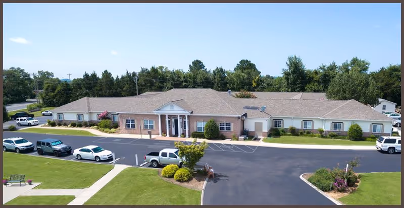 Front exterior of a single-story brick senior living building with a central entrance, parking lot, landscaped lawns, and several parked cars.