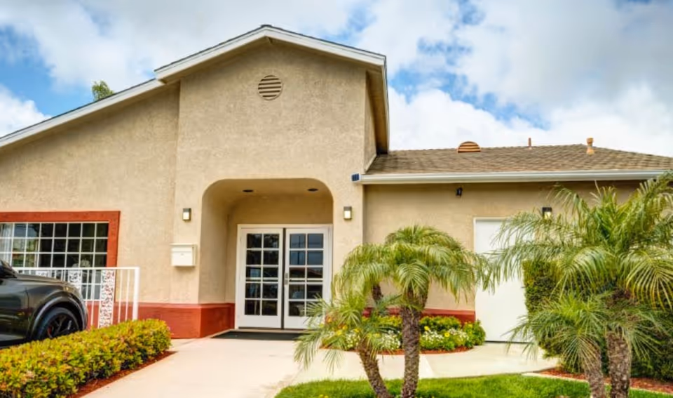 Beige single-story memory care facility with double glass doors and palm trees at entrance.