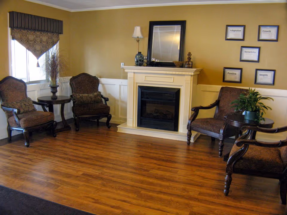 A cozy sitting area with four upholstered armchairs arranged around a white electric fireplace. There are two small round wooden tables with decorative plants and a lamp on the fireplace mantel. The walls are painted yellow with white wainscoting, and framed inspirational words hang on the wall. The floor is wood with a warm finish.