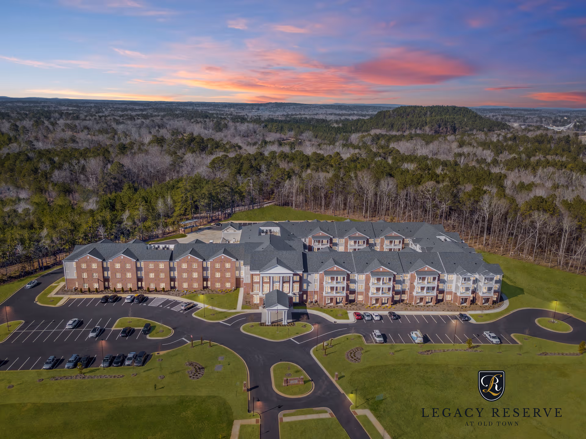 Aerial view of a large brick senior living building with parking lots, lawns, and surrounding woods under a colorful sky.