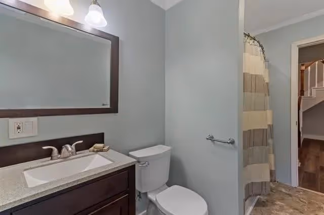 A bathroom with a sink and countertop on the left, a mirror with two light fixtures above it, a toilet next to the sink, and a shower with a striped curtain on the right. The bathroom walls are painted light gray, and there is a doorway leading to a hallway with wooden flooring.