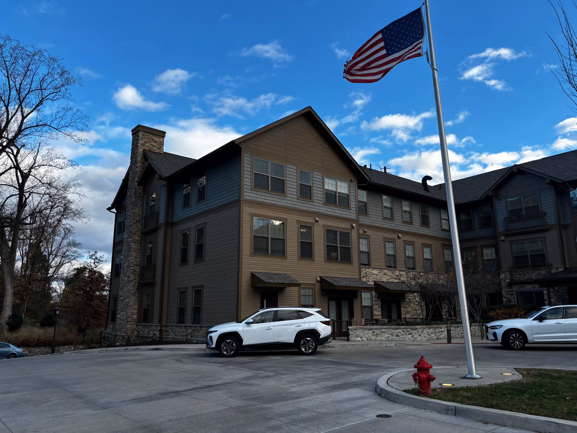 Exterior view of a senior living facility building with a stone chimney, multiple windows, and a brown and gray facade. Two white cars are parked in front on a concrete driveway. An American flag on a tall flagpole and a red fire hydrant are visible in the foreground under a partly cloudy blue sky.