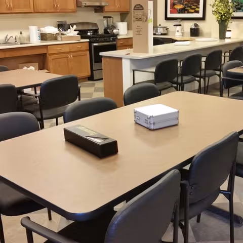 A communal dining area with tables and chairs, adjacent to a kitchen space featuring wooden cabinets, a stove, and various kitchen appliances. The tables have some items placed on them, and there are framed pictures and a plant on the wall in the background.
